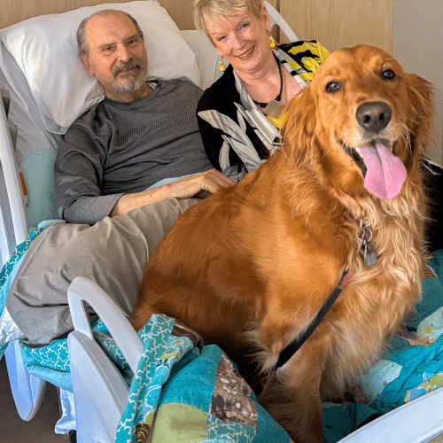 A photo of Cindy and Larry at Hospice Quinte. Their dog Lady, a golden retriever, sits on the hospital bed with them. They are all smiling for the photo.