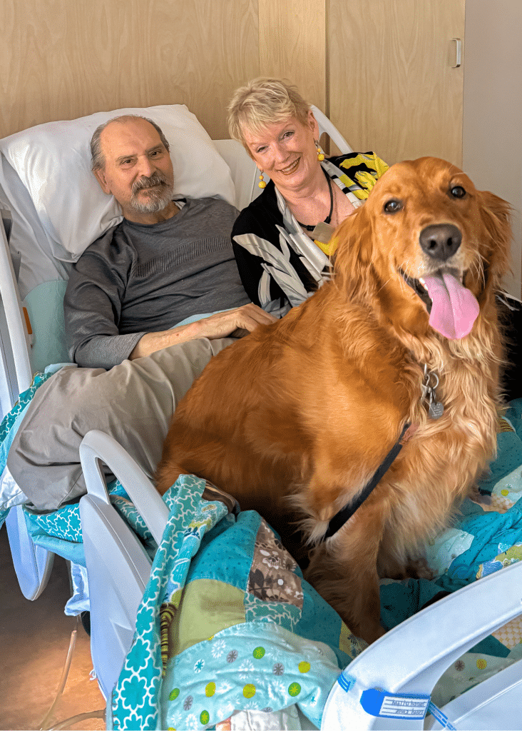A photo of Cindy and Larry at Hospice Quinte. Their dog Lady, a golden retriever, sits on the hospital bed with them. They are all smiling for the photo.